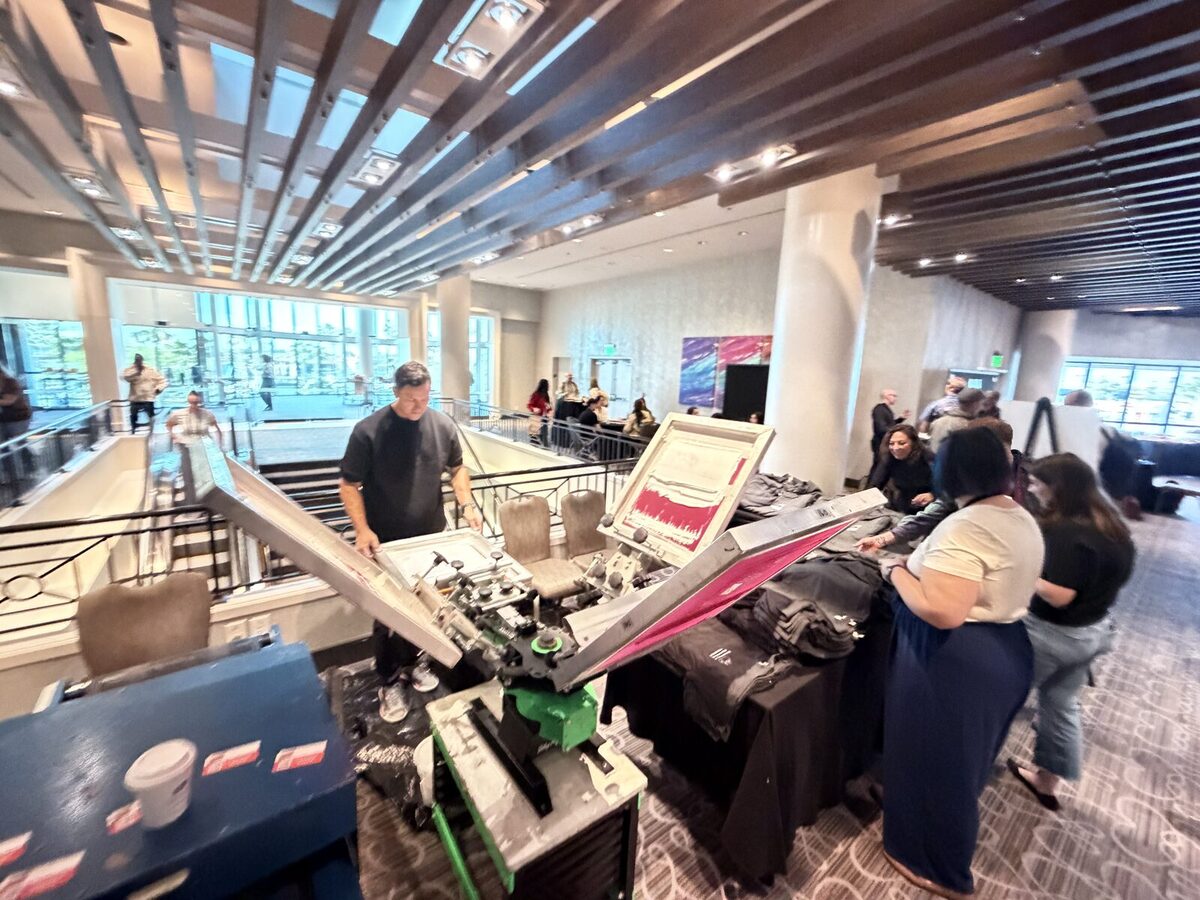 An operator running an automatic press as attendees line up at a merch booth
